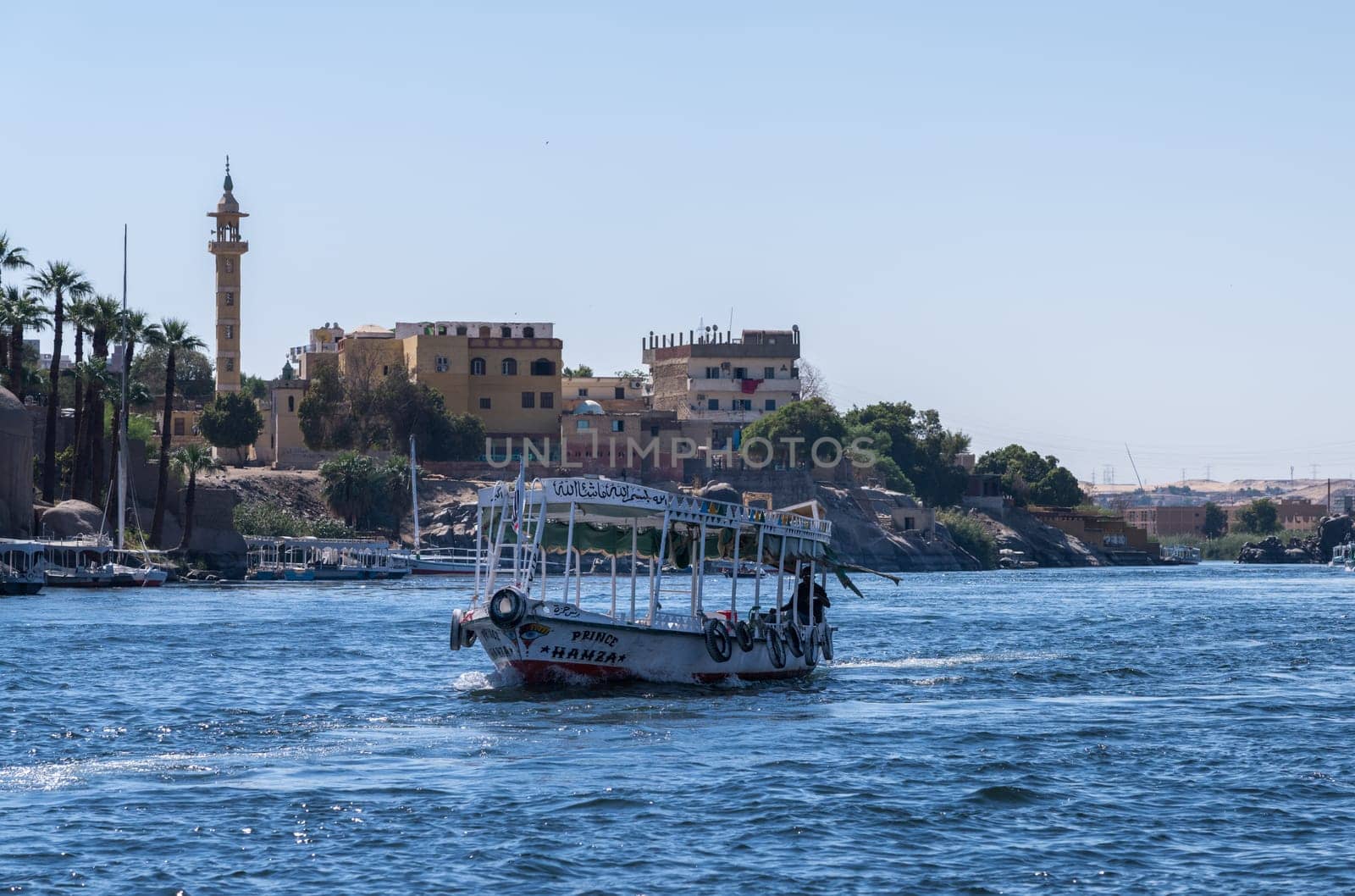 Aswan, Egypt - 4 November 2024: Ferry boat approaches the dock on the Nile with mosque and homes on the shore