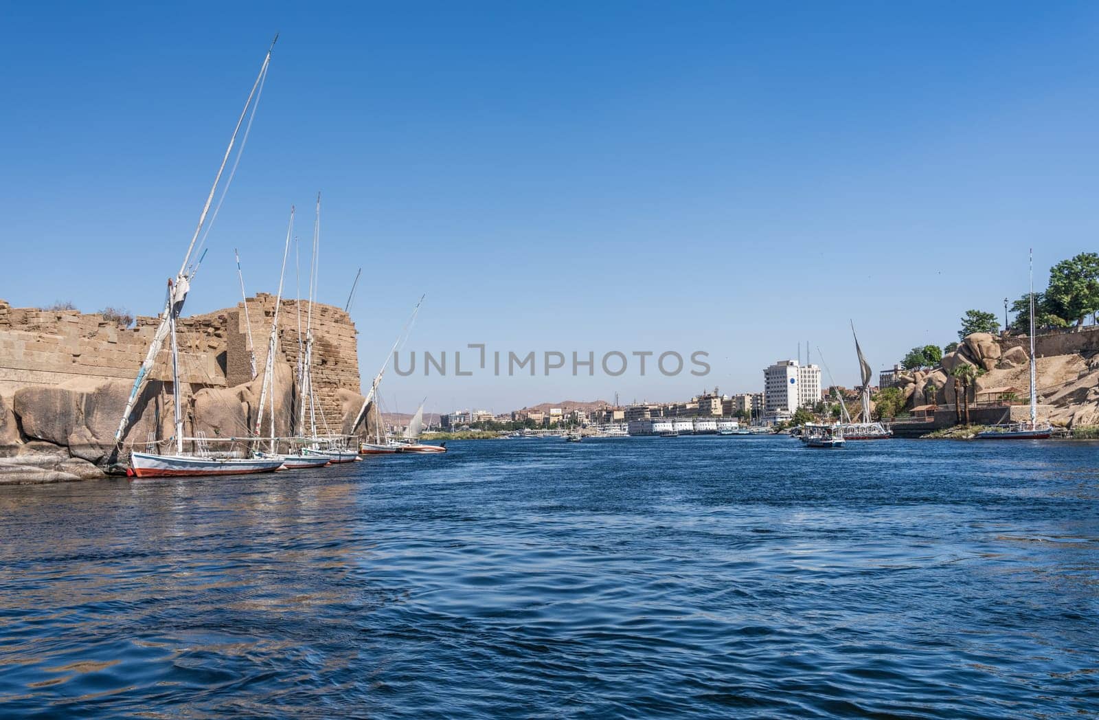Skyline of Aswan in distance with Felucca boat docked by Ruins of Abu and many river cruise boats docked
