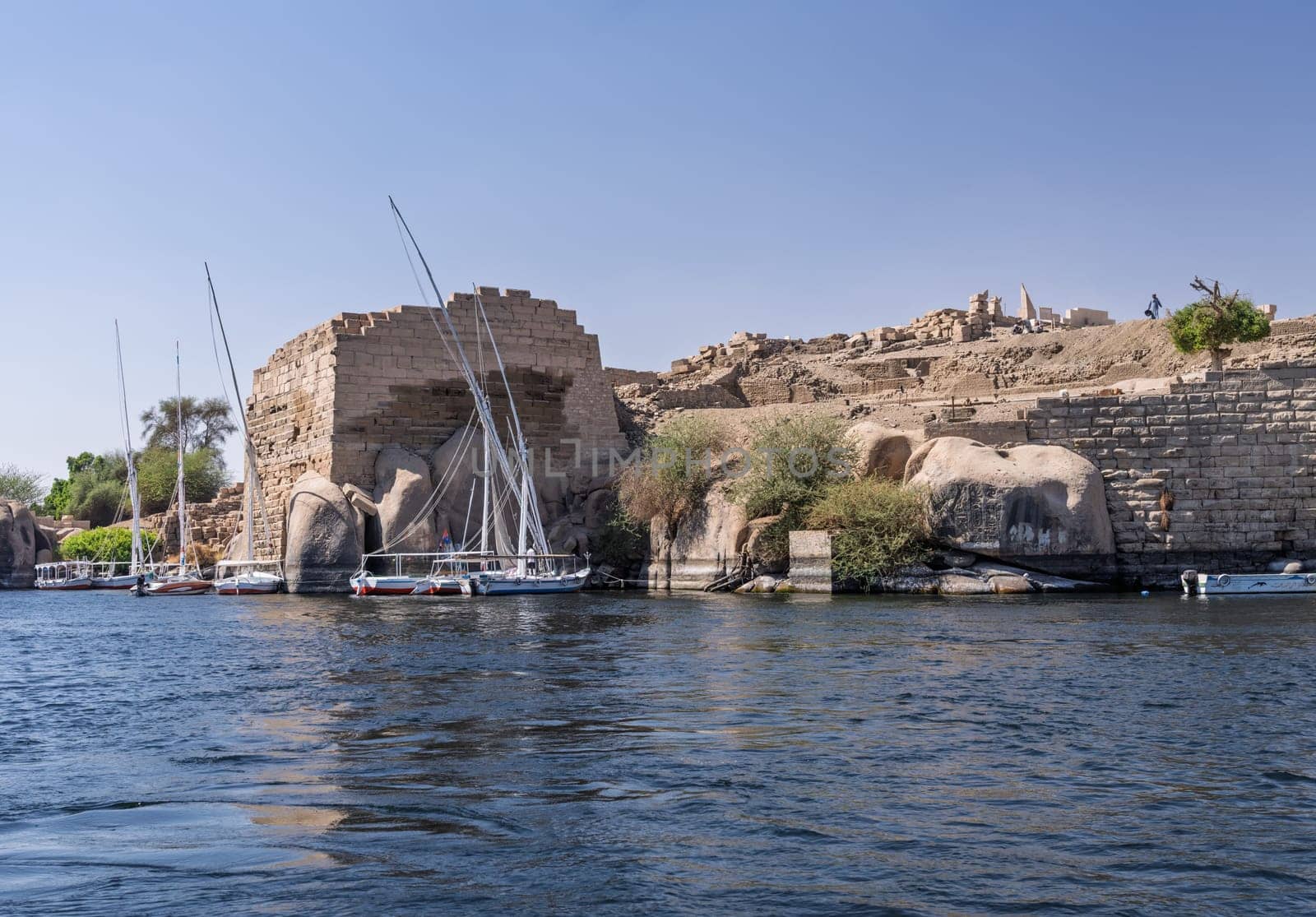 Felucca boat docked by Ruins of Abu outside Aswan in Egypt and other river cruise boats docked by the massive stone walls