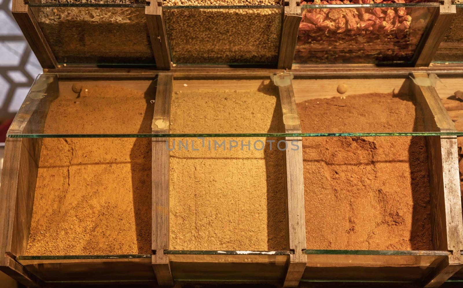 Wooden display of various spices in a market, with glass partitions and overhead lighting, showcasing flavors
