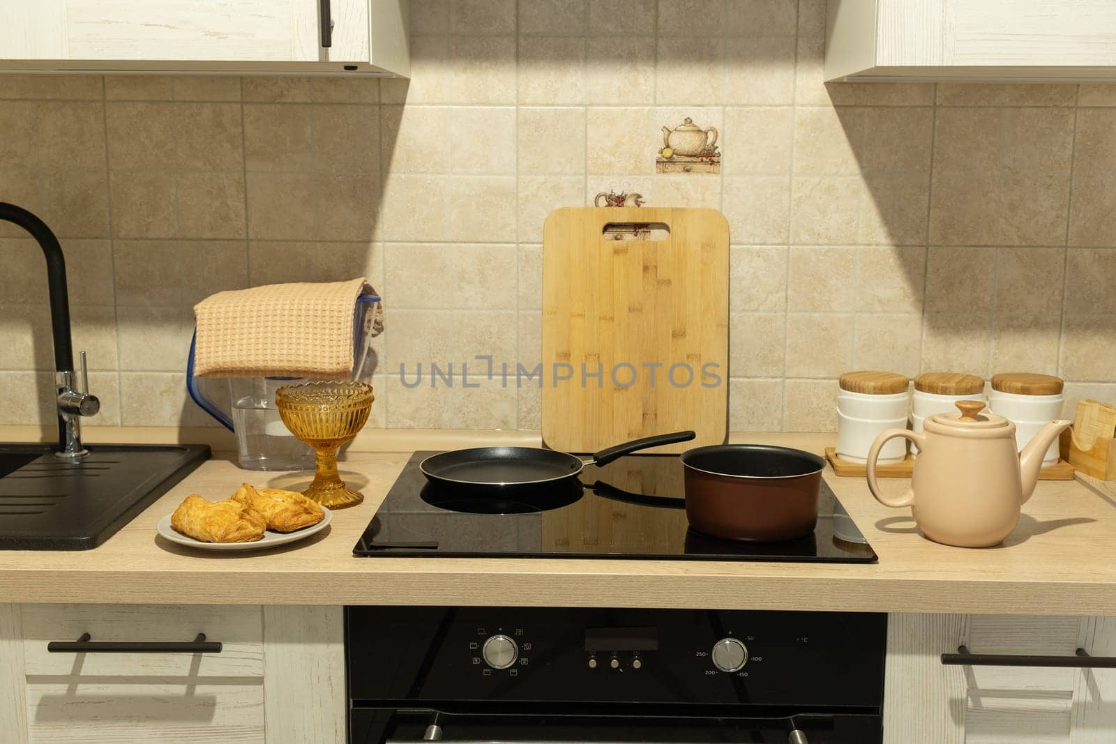 Cozy kitchen setup featuring a teapot, buns on a plate, and cookware on a stovetop. Tiled backsplash, cutting board, and utensils complete the organized space