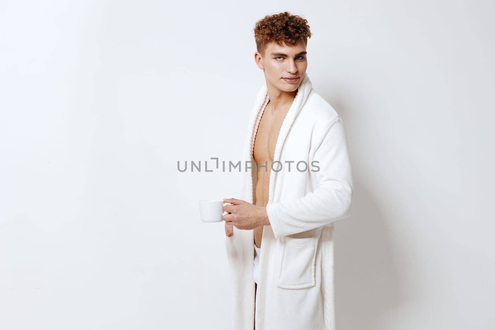 Confident young male in a cozy white robe enjoying a cup of coffee, showcasing a relaxed and trendy lifestyle in a minimalist indoor environment.