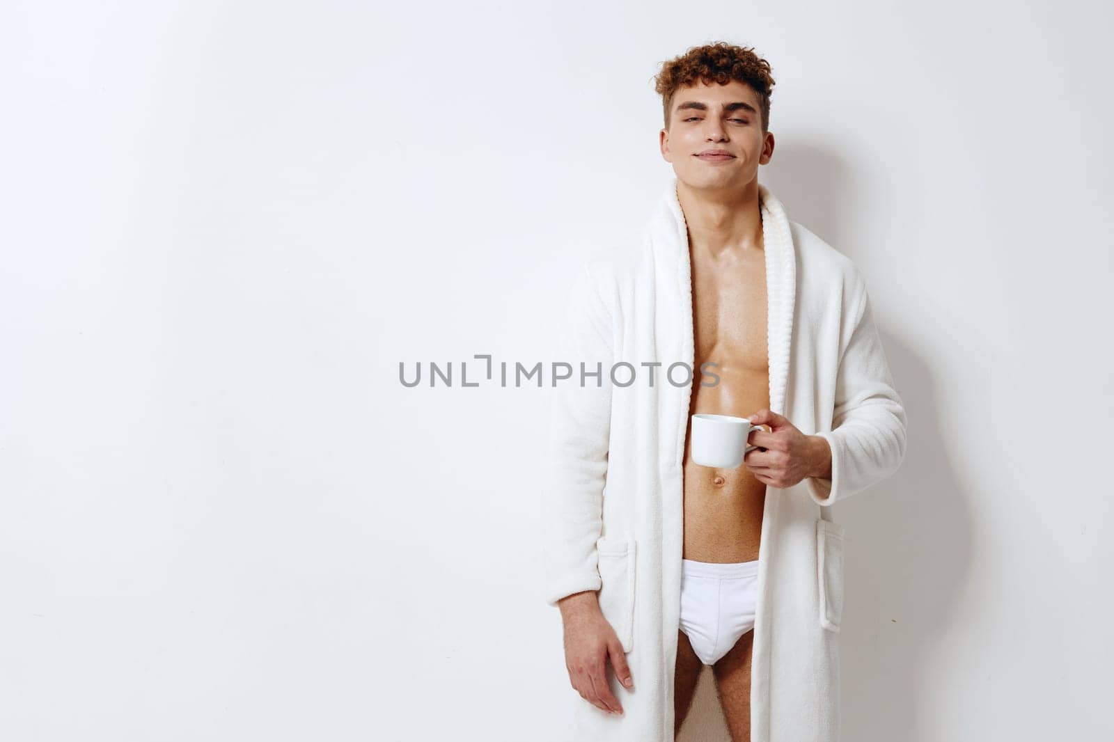 Smiling young man wearing a fluffy white robe, enjoying a cup of coffee. Emphasizing relaxation and comfort in a peaceful indoor setting.