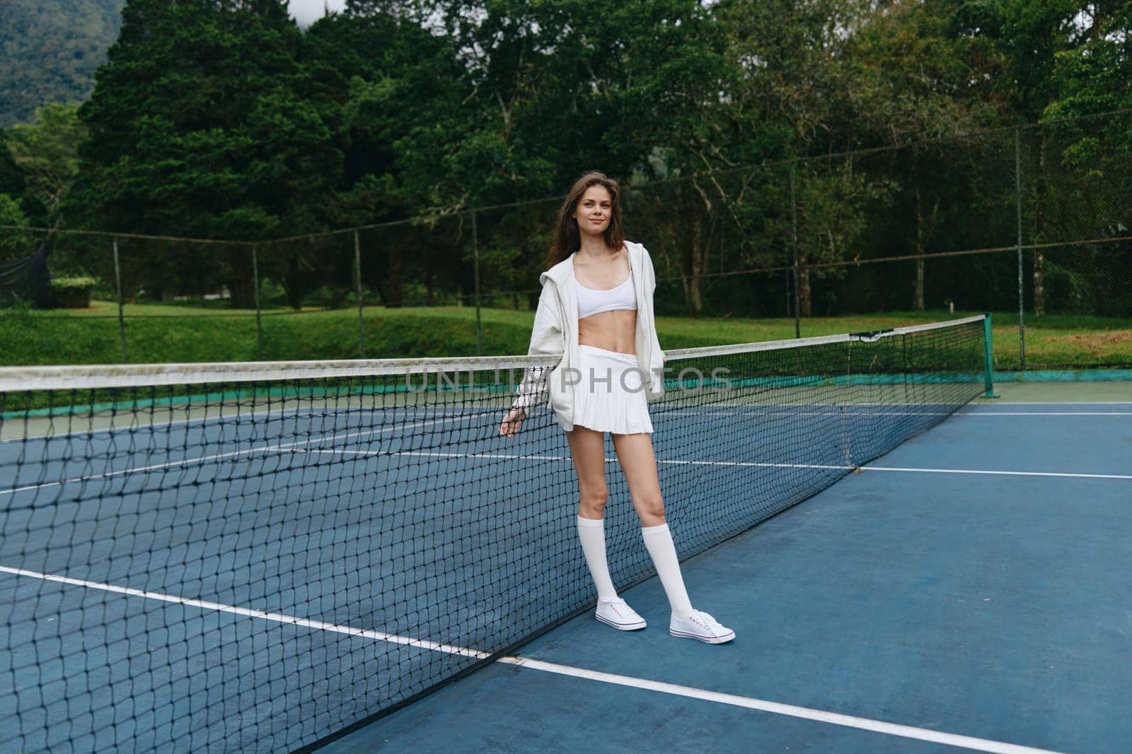 A confident young female tennis player dressed in white sportswear stands on a tennis court, exuding enthusiasm and joy The lush green background enhances the vibrant outdoor atmosphere, perfect for a summer sports concept