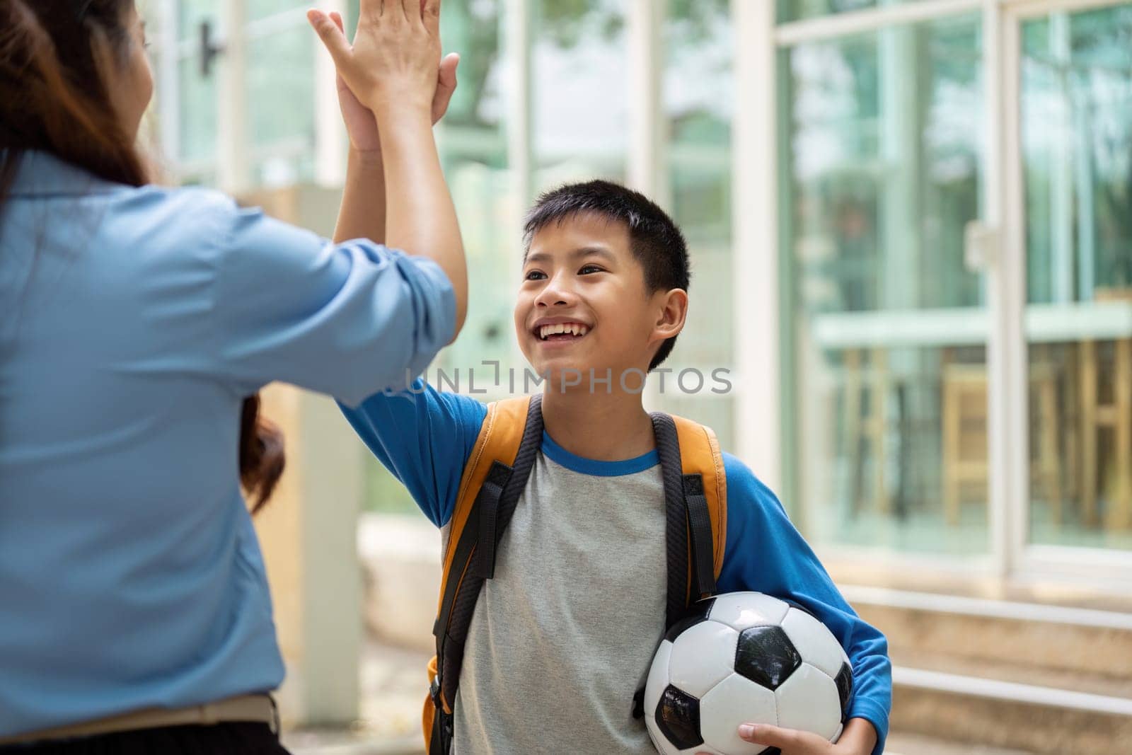 A joyful boy high-fives his teacher outside a school, excited about returning to classes and playing soccer. This captures the spirit of enthusiasm for learning and sports.