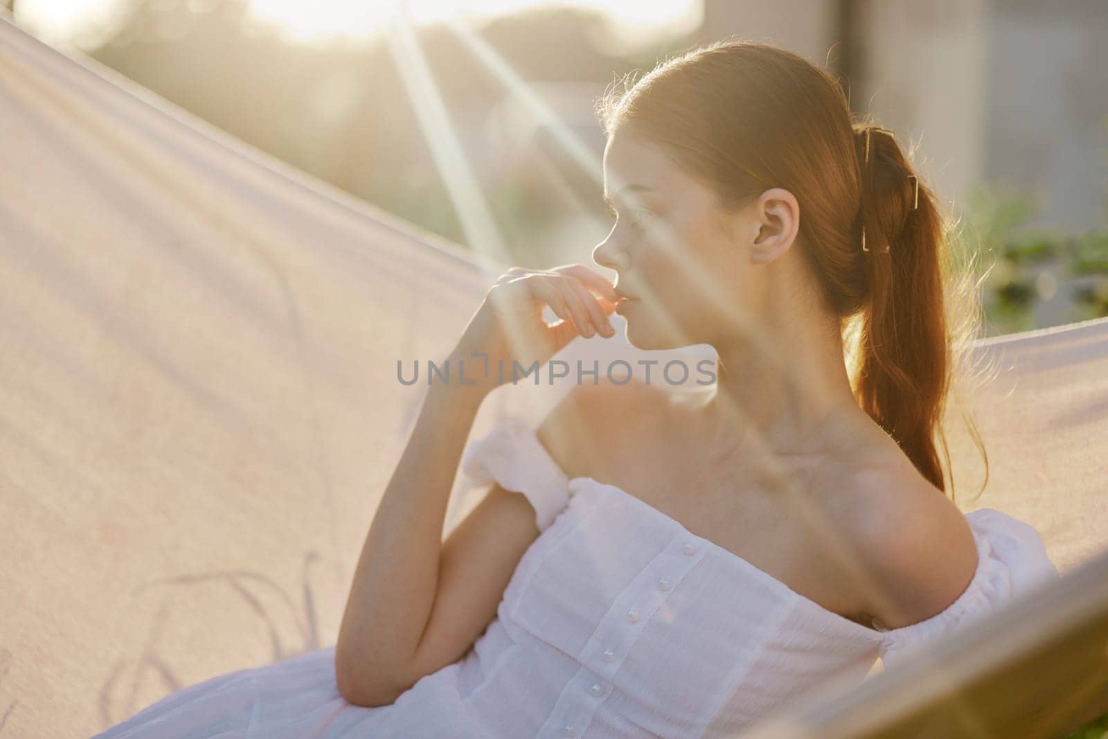 A young woman in a flowing white dress relaxes in a hammock, bathed in warm sunlight, radiating calm and serenity, ideal for themes of leisure and summer relaxation