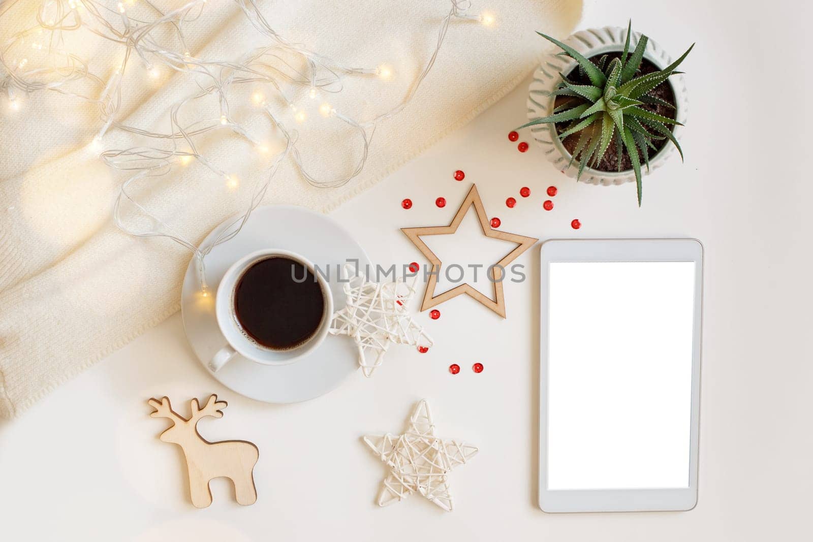 Christmas decorations and a digital tablet with a blank screen, lies on a white table with a glowing garland, a deer, stars, snowflakes, a white cup of coffee. View from above.