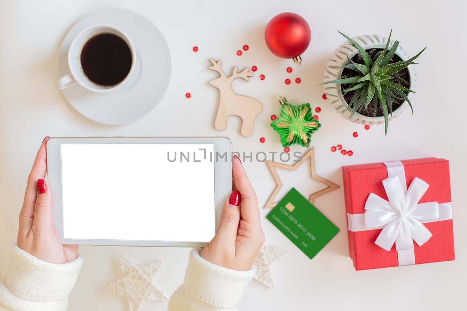 The hands of a woman are holding a digital tablet, a blank credit card, a red gift box , a cup of coffee and a succulent plant , Christmas decor and toys are lying next to the white table. Mock up