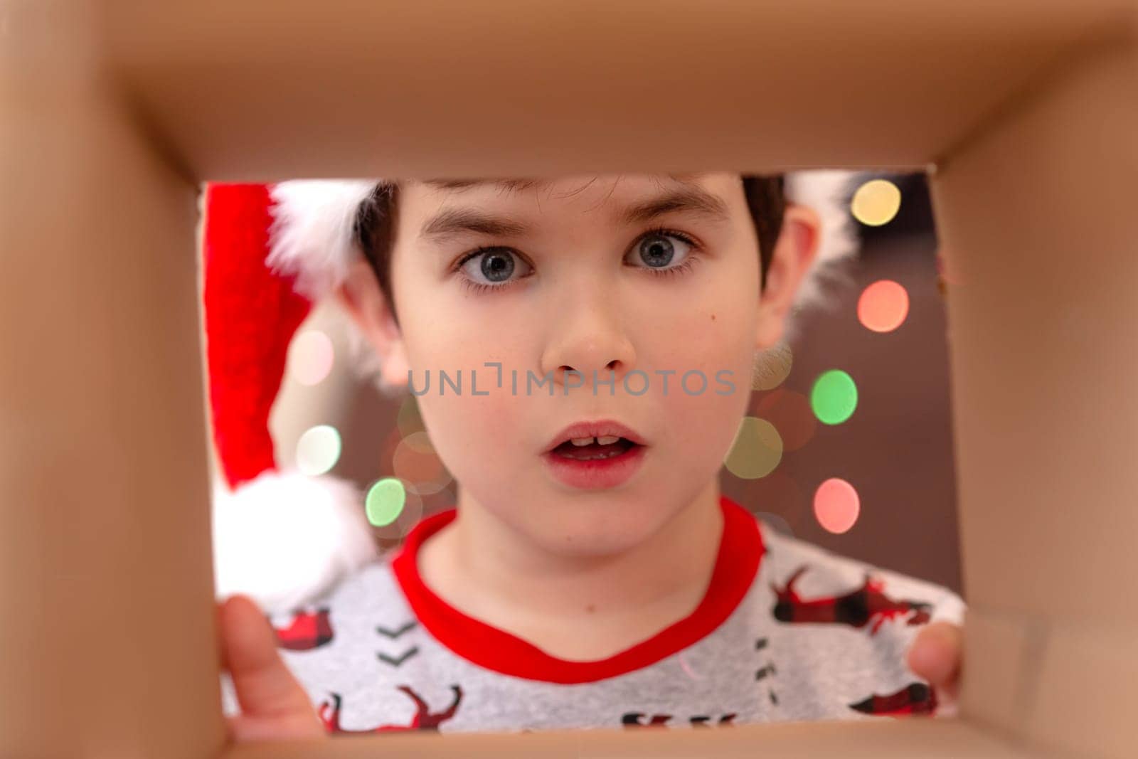 The shock of emotion in a boy in a New Year's jumper and a hat of Santa Claus looks into a box with a gift, a view from the inside, against a background of bokeh from light garlands.