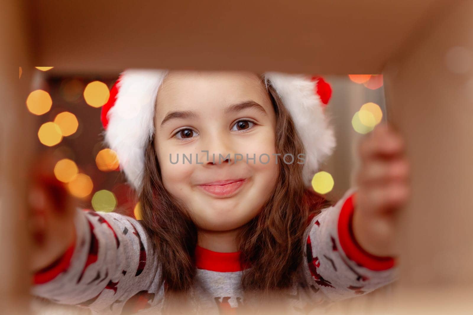A happy girl in a New Year's jumper and Santa Claus hat takes a present out of the box, holding out his hands to the present, on the background of bokeh of lights garlands