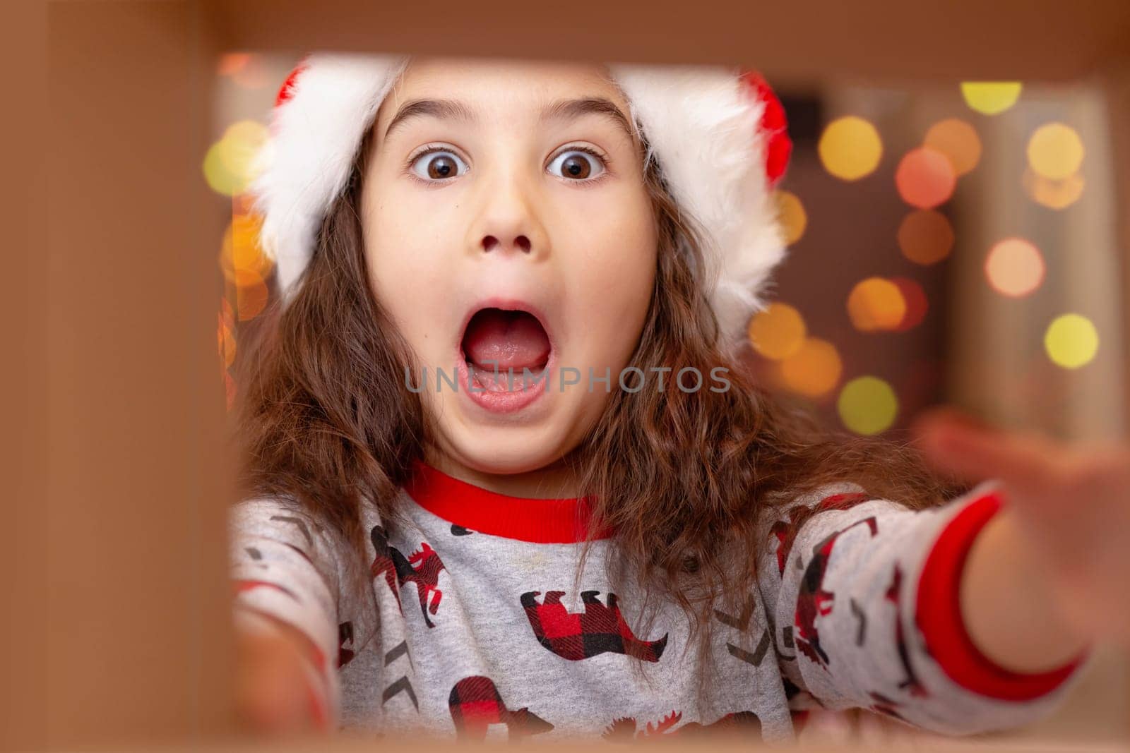 Close-up portrait. A joyful, surprised, beautiful girl in a New Year's jumper and a Santa Claus hat takes a gift out of the box, stretching out her hands to the gift, view from the inside.