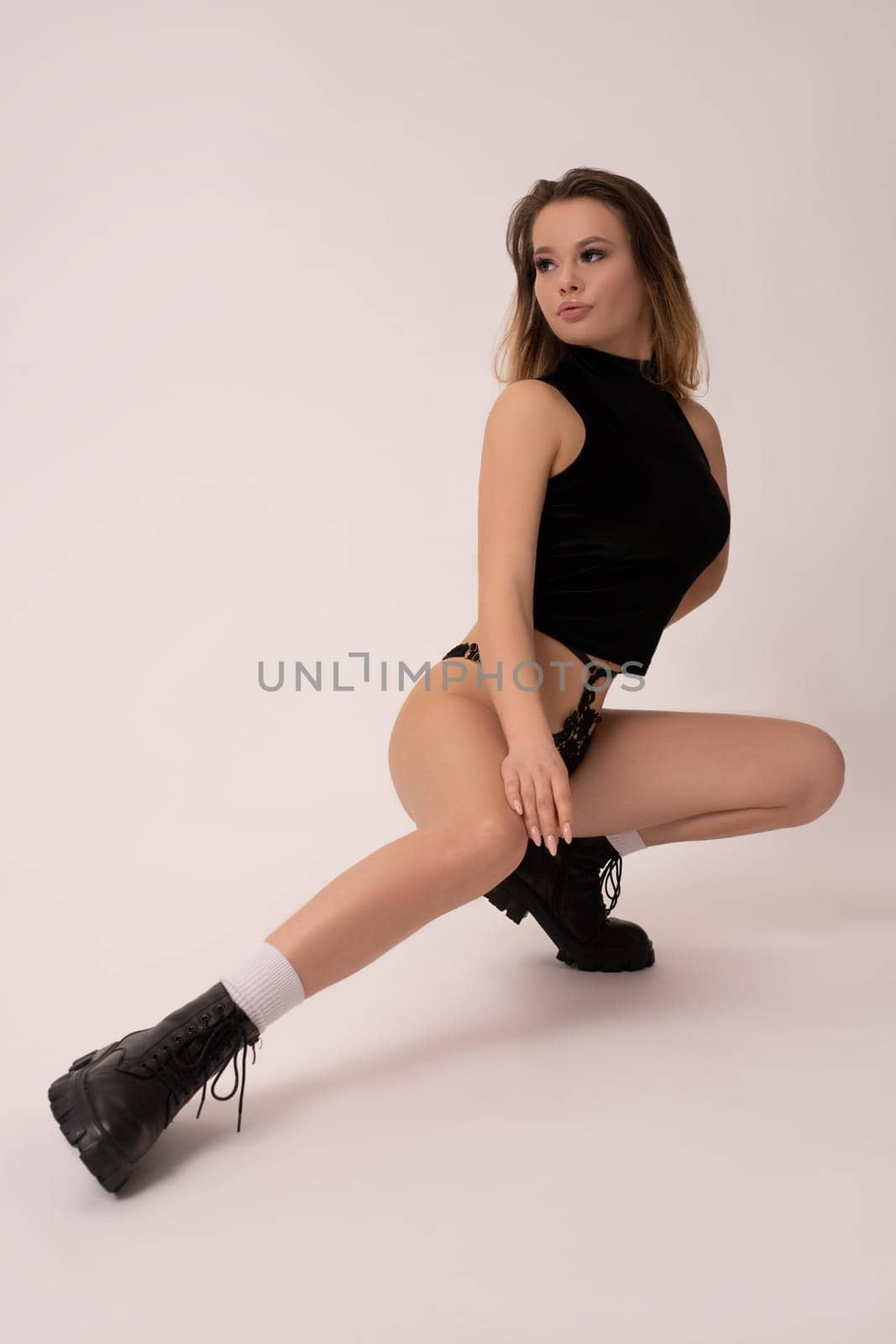 Full body of sensual slim woman in black lingerie and boots sitting on haunches and looking away against gray background