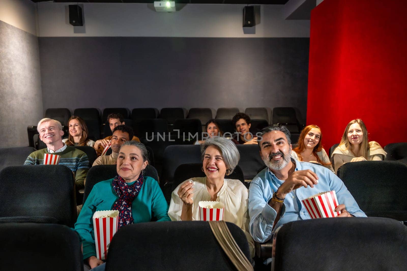 People of different ages and ethnicities are watching a film and eating popcorn in a movie theater, experiencing a shared moment of entertainment