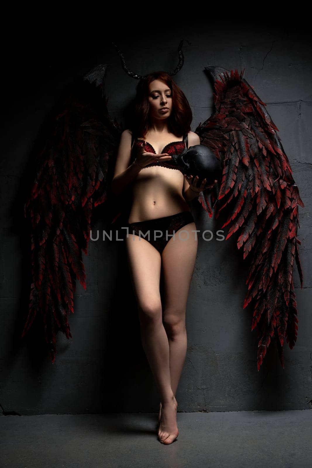 Full body of seductive female with horns and black wings looking at camera on black background with skull