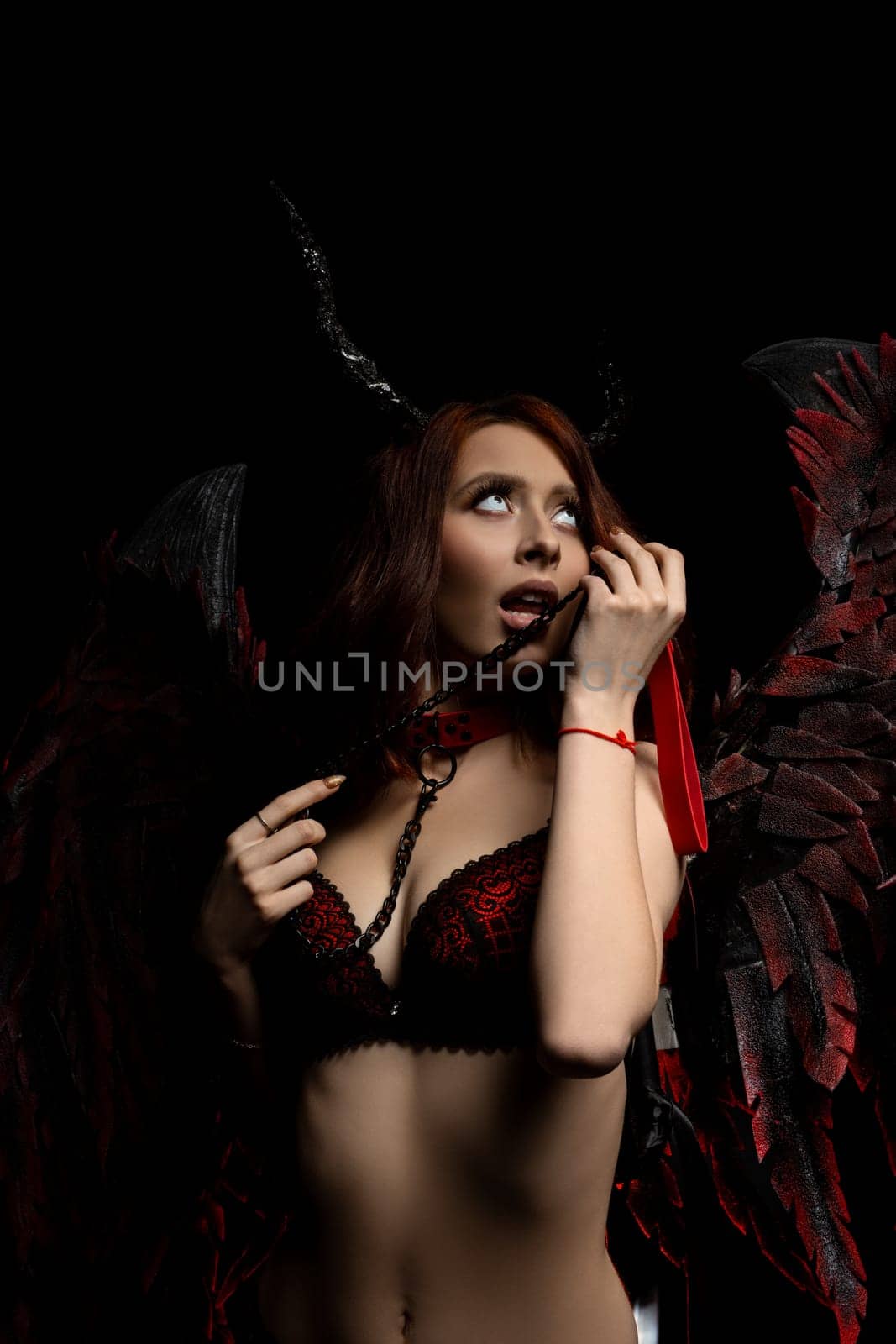 Seductive female in underwear with horns and wings looking up while touching neck collar on black background in dark studio