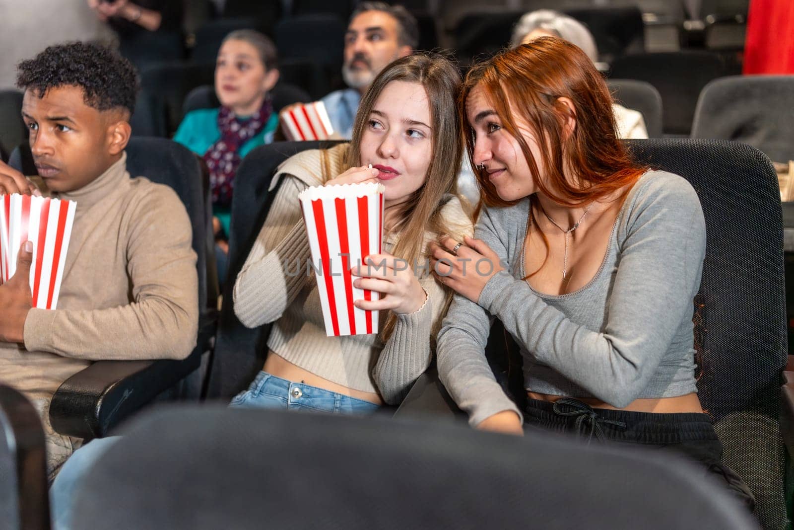 Two friends are whispering and eating popcorn while watching a movie at the cinema, surrounded by other people
