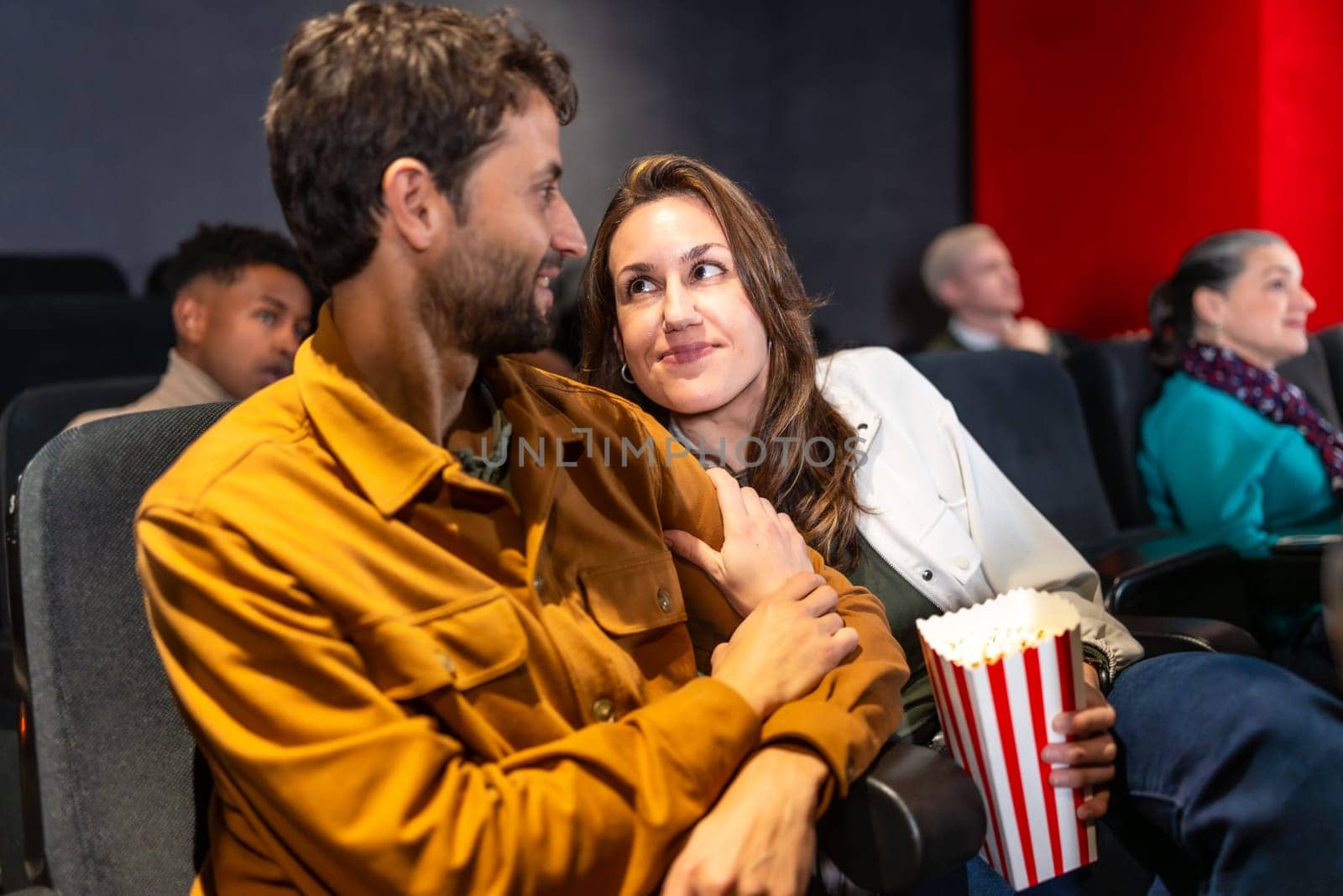 Couple enjoying a cozy movie night, sharing popcorn and affection while surrounded by a lively audience in a bustling cinema