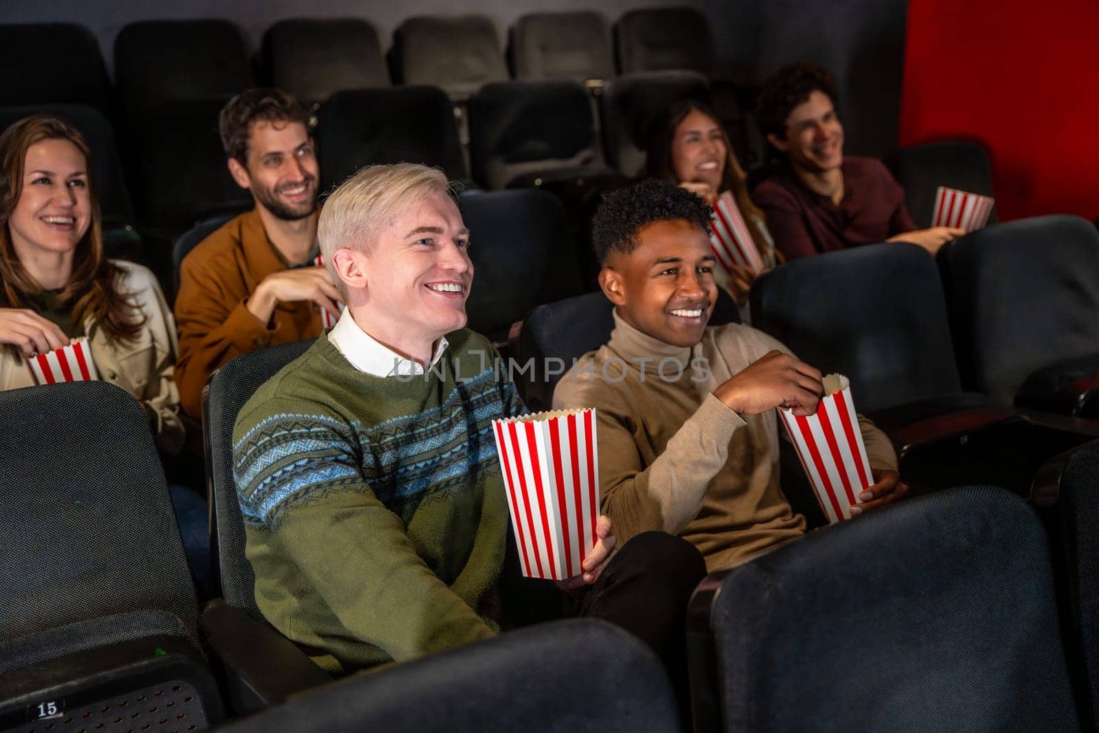 Diverse group of friends laughing and eating popcorn while watching a movie at the cinema