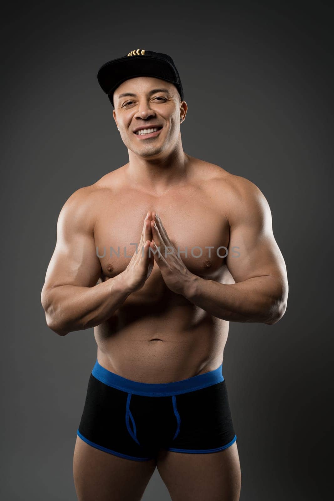 Full body of sexy shirtless trainer in underpants and cap showing muscles and looking at camera on gray background in studio