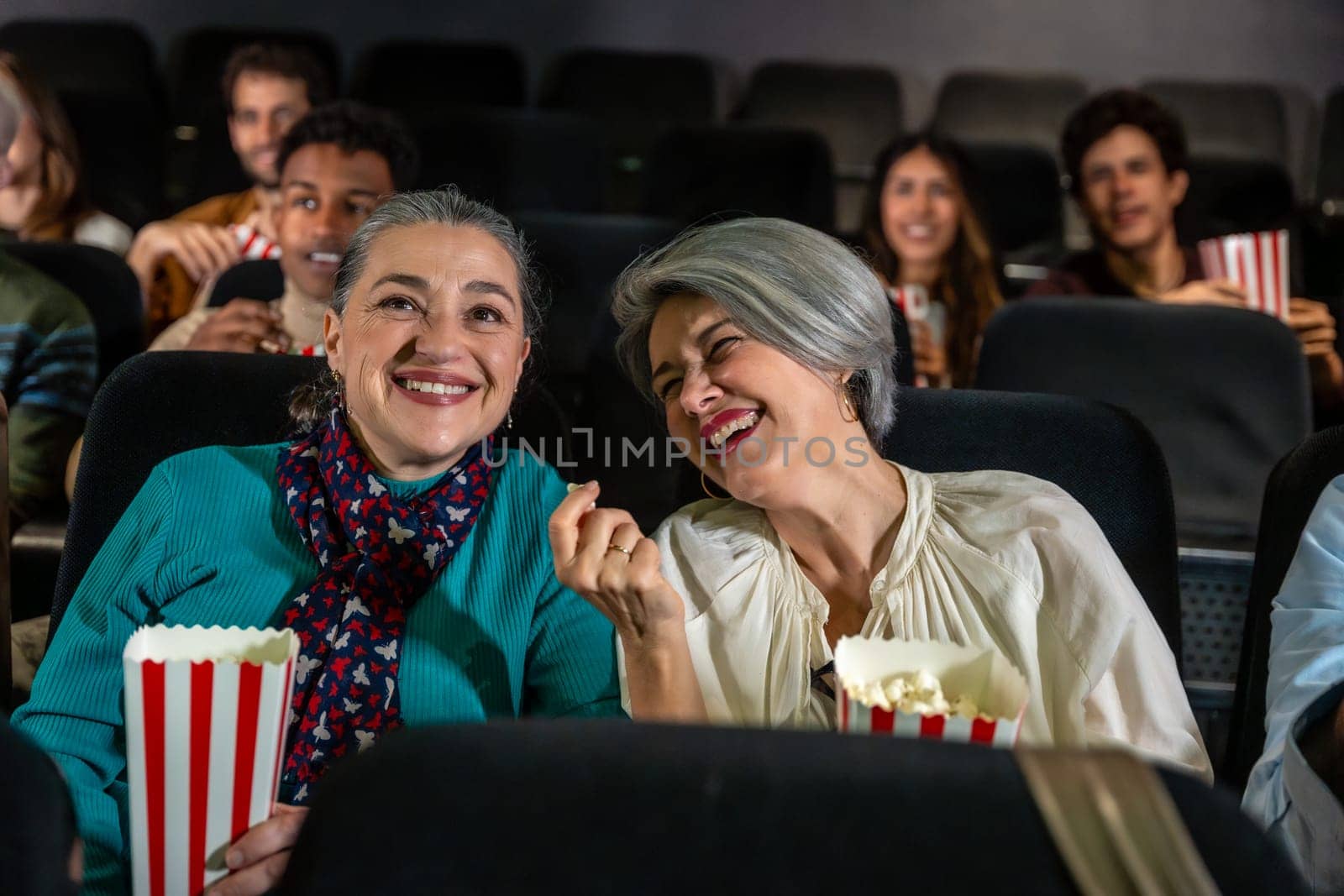 Two cheerful senior women laughing and eating popcorn while watching a movie at the cinema, sharing a joyful moment together
