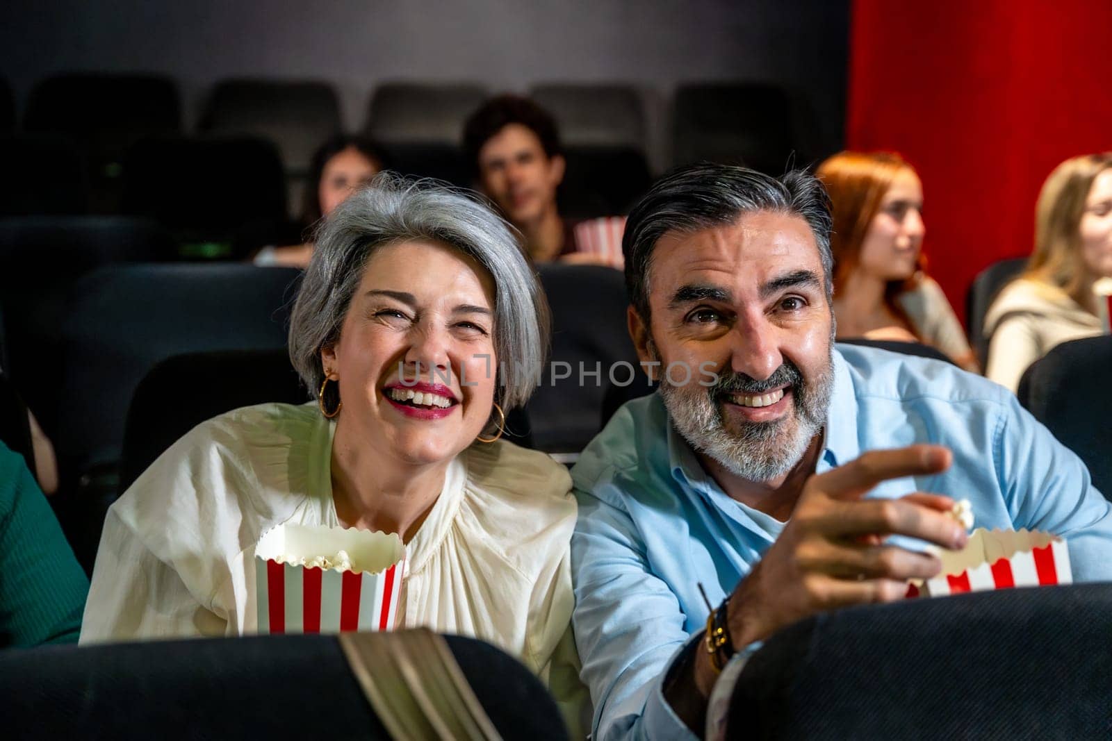 Happy mature couple laughing and having fun watching a movie at the cinema, eating popcorn
