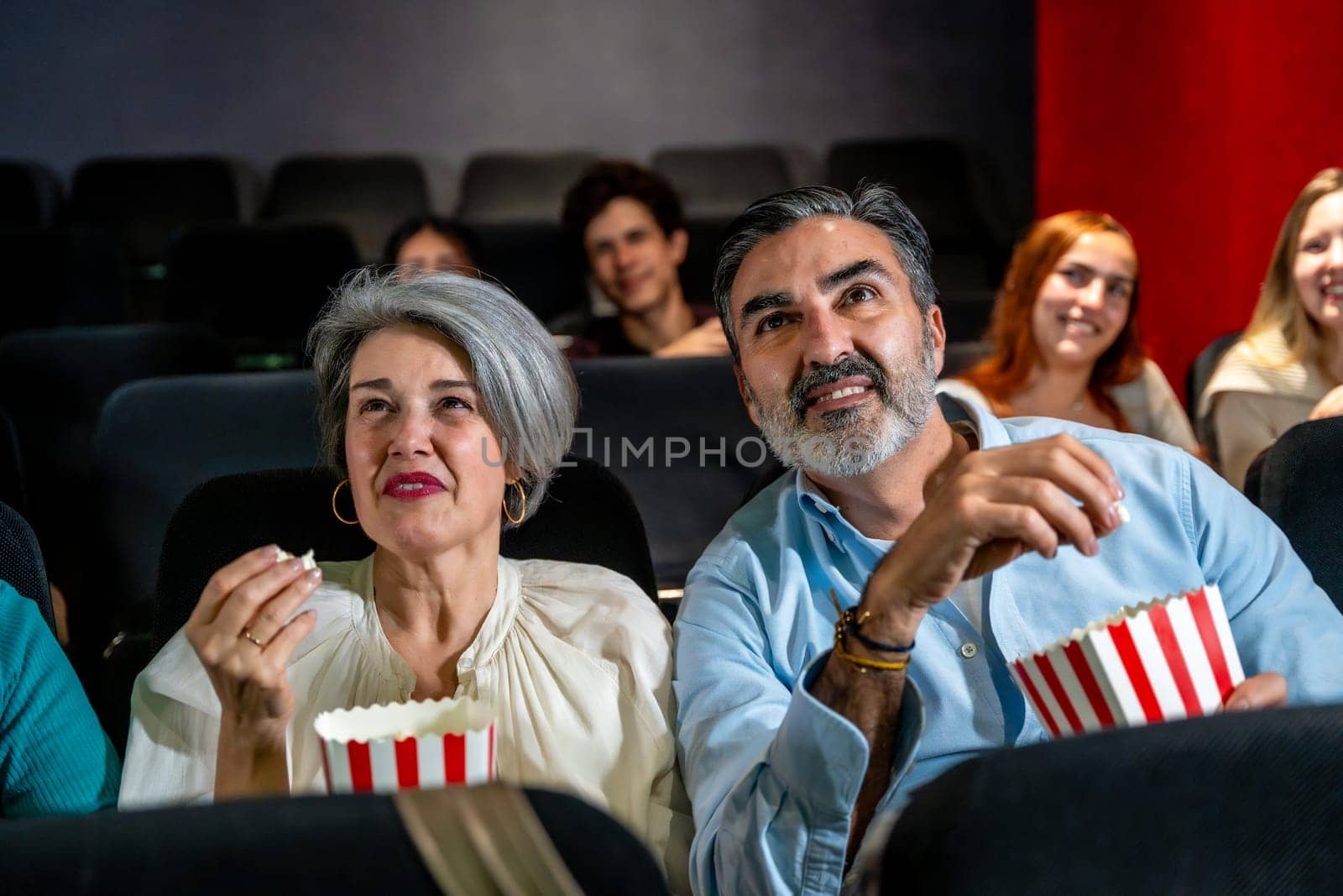 Senior couple watching a movie and eating popcorn, enjoying a night out at the cinema with other people