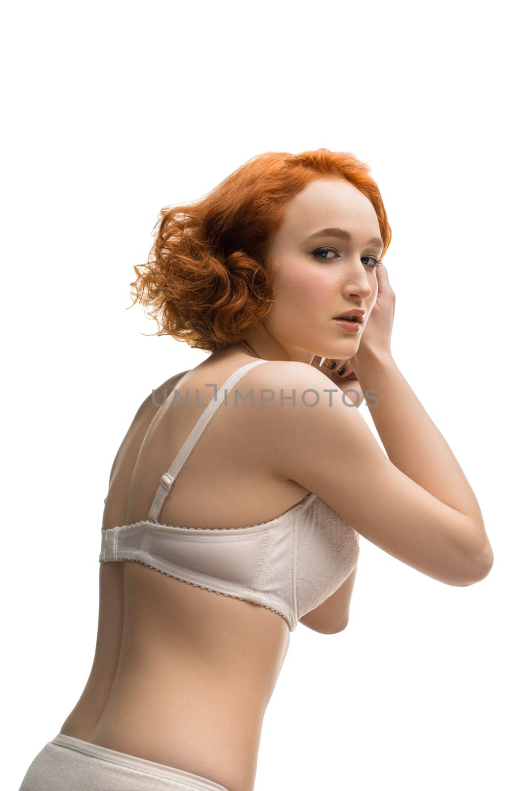 Side view of alluring female with curly red hair wearing white lingerie standing in studio against white background and looking at camera
