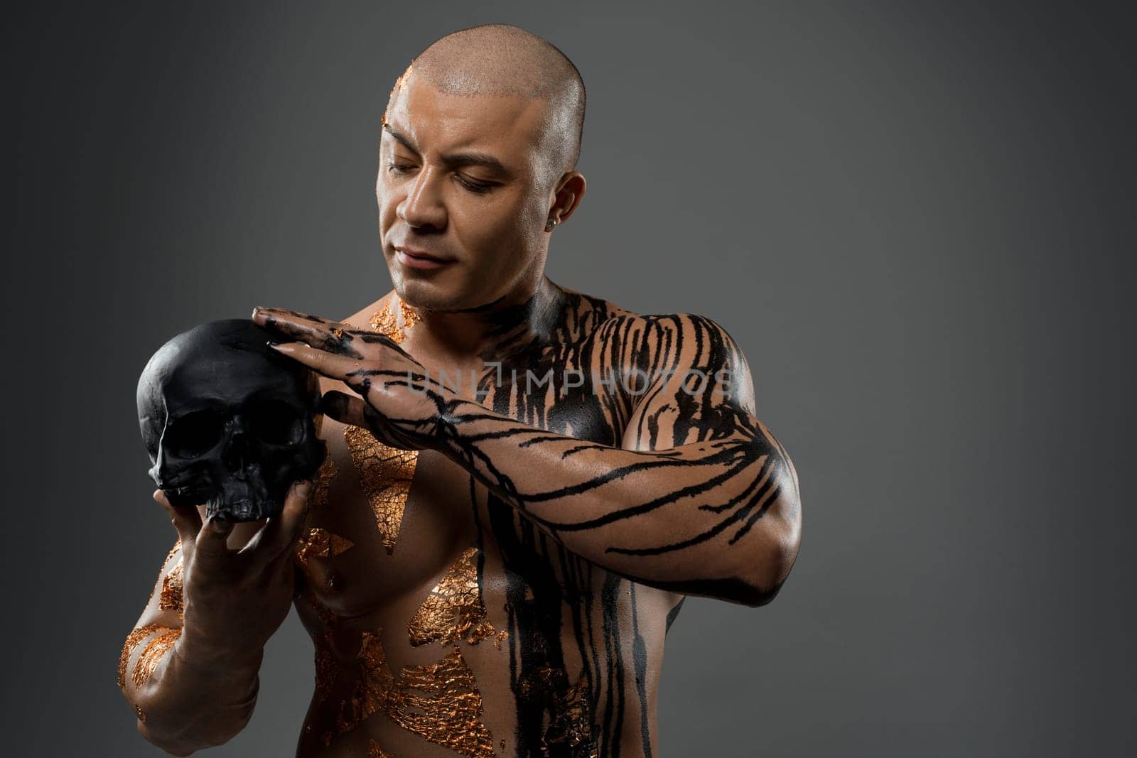 Handsome sexy male with flowing paint stains on fit naked torso touching black skull on gray background in studio