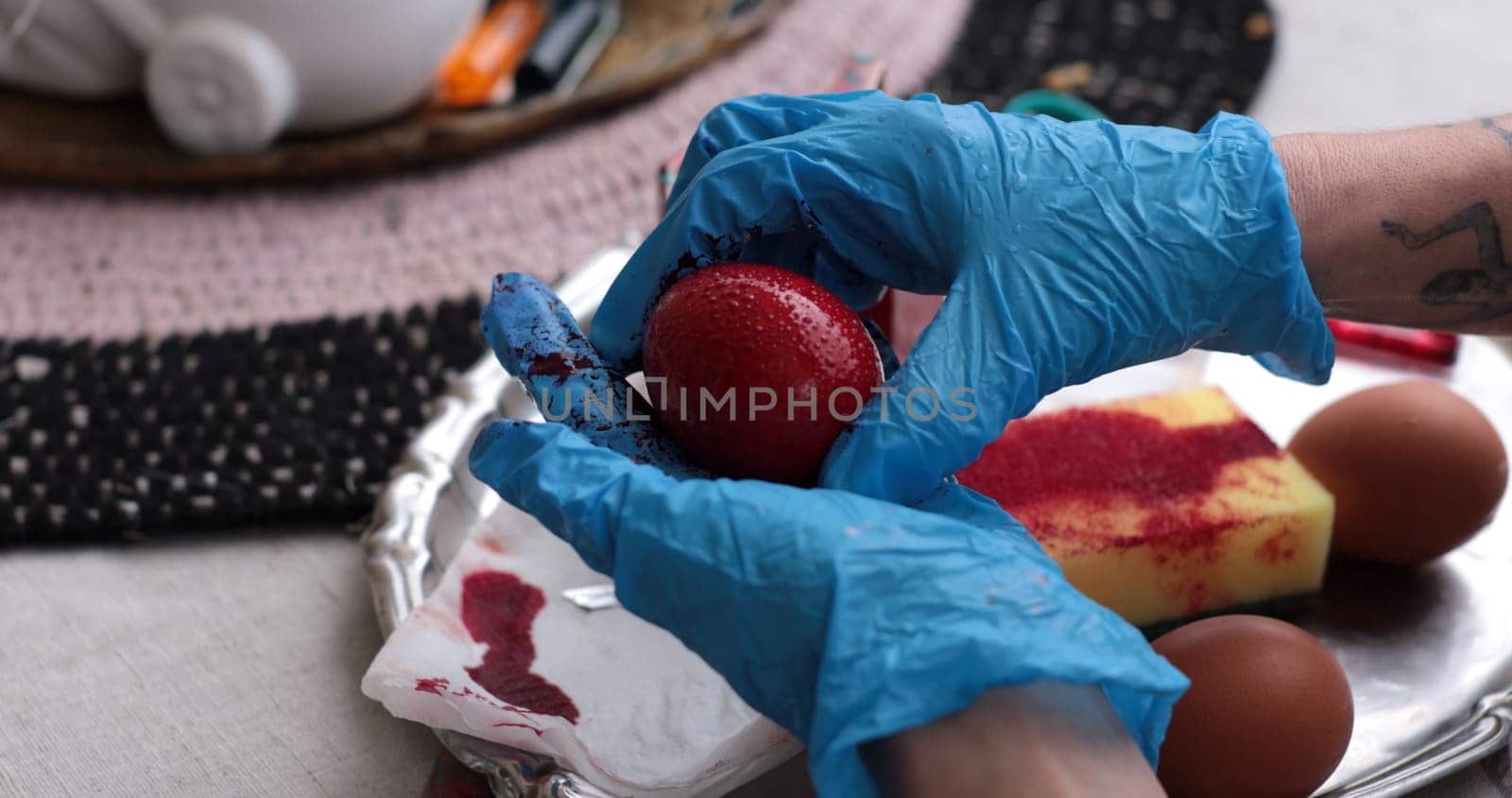 Hands wearing blue gloves hold a red Easter egg being painted as part of a coloring activity. Surrounding items include brown eggs and dye, hinting at the joy of spring celebrations.