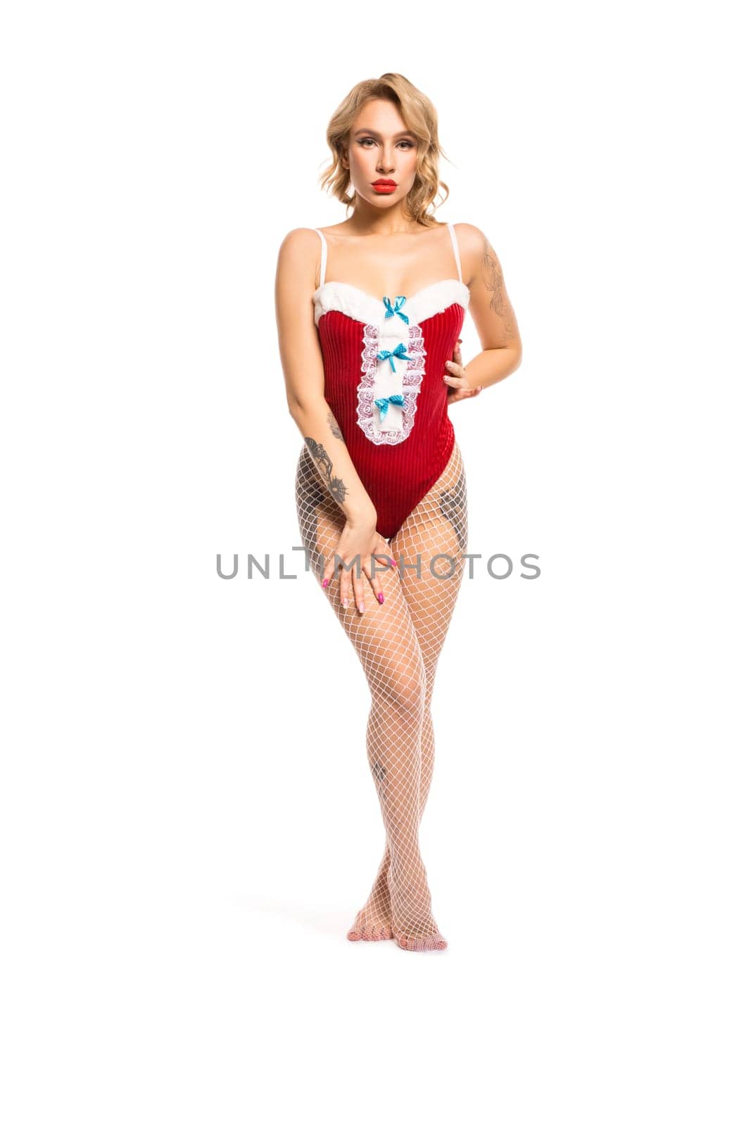 Full body of graceful tempting female in red Santa bodysuit and net tights standing with hand on waist against white background and looking at camera