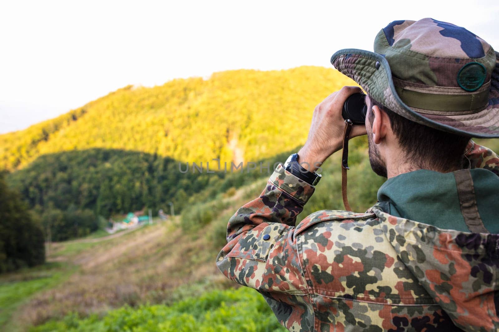 Soldier using binoculars in mountain forest is observing enemy positions