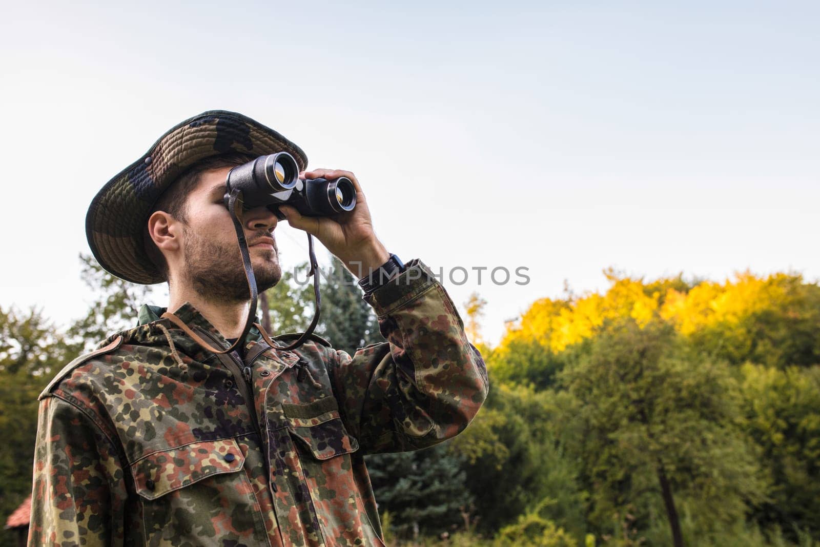 Soldier using binoculars while observing enemy positions in forest during military operation