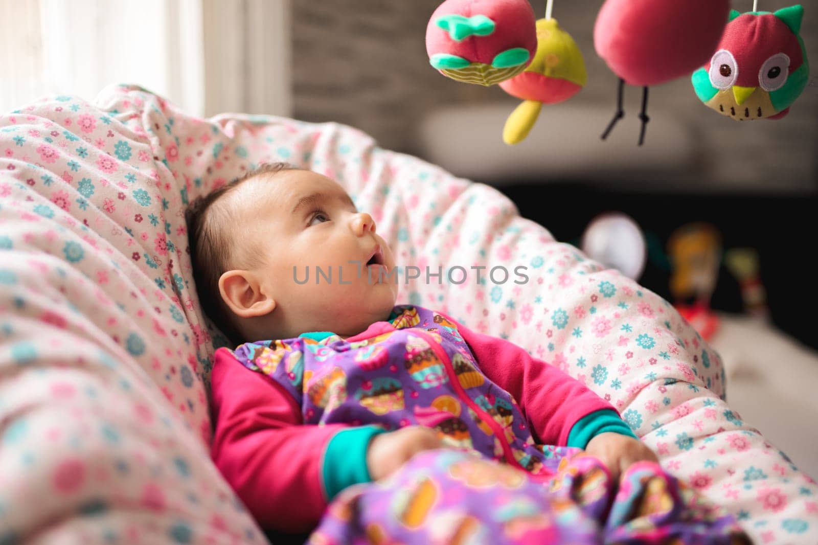 Newborn baby girl in a crib looking at colorful toys hanging above.