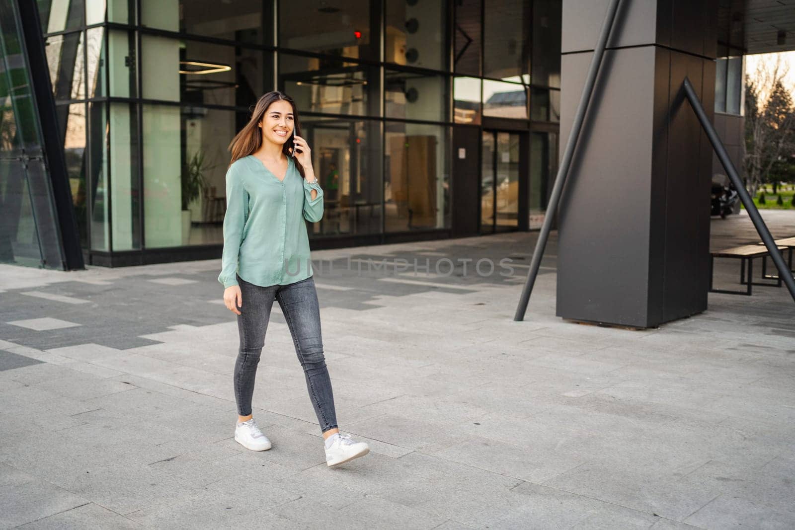 Smiling young brunette girl walks in outdoors and makes a phone call.