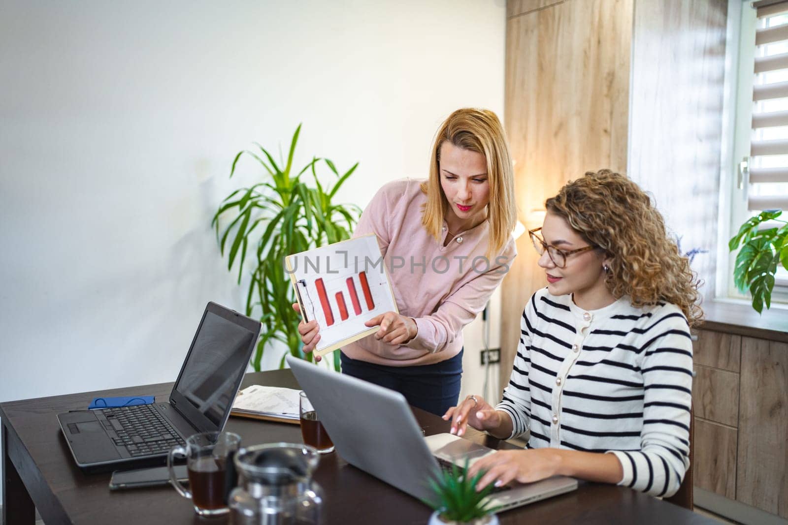 Two female colleagues are working from home, one is sitting at a desk using a laptop, while the other is standing next to her with a chart in hand, working together on a business report.
