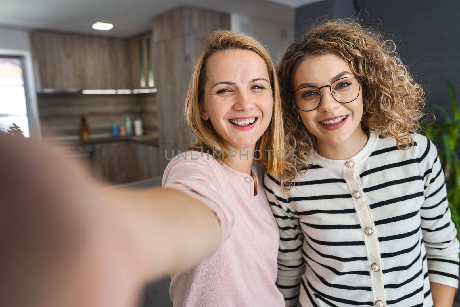Two women are standing in front of a camera, holding a phone up, and smiling as they take a selfie together.