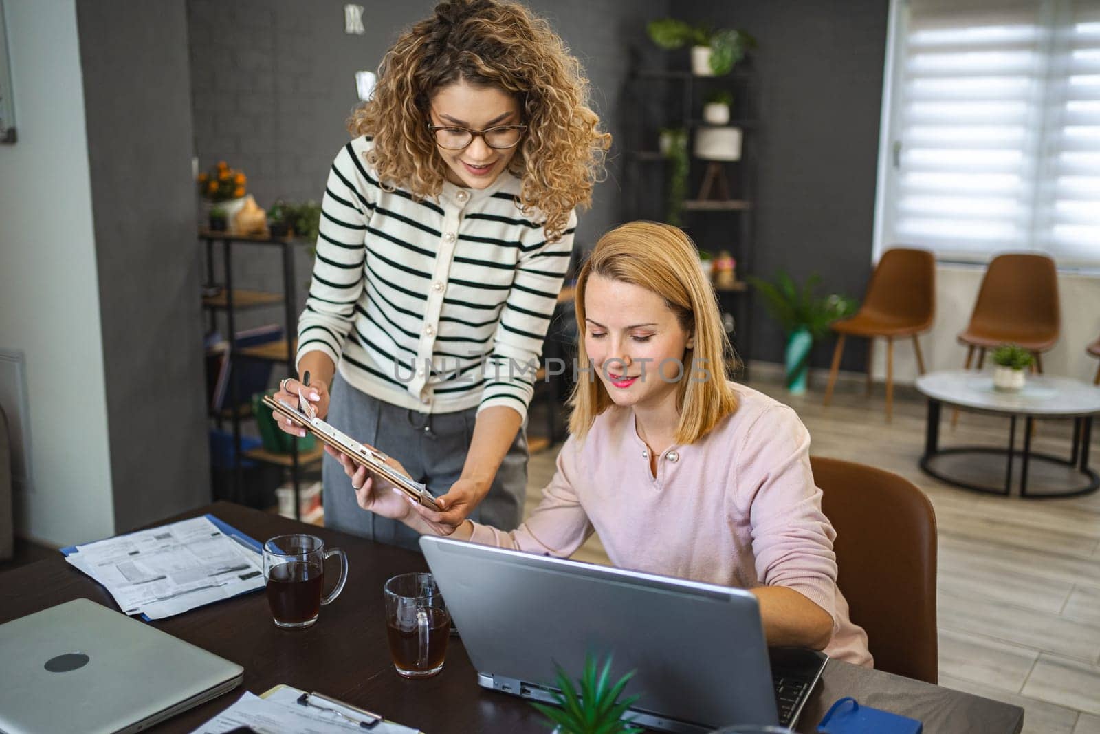 Two female colleagues are working from home, one is sitting at a desk using a laptop, while the other is standing next to her with a document in hand, working together on a business report.