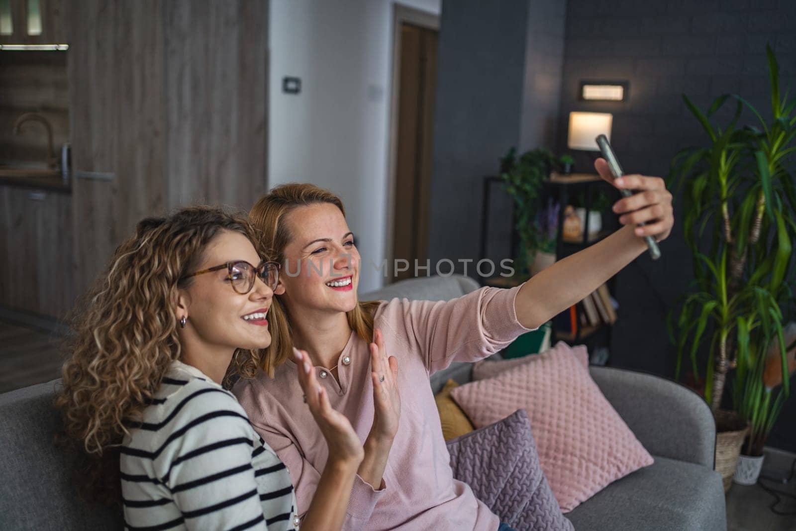 Two female friends are sitting on the couch in the living room and making a video call and sending greetings to the interlocutor.