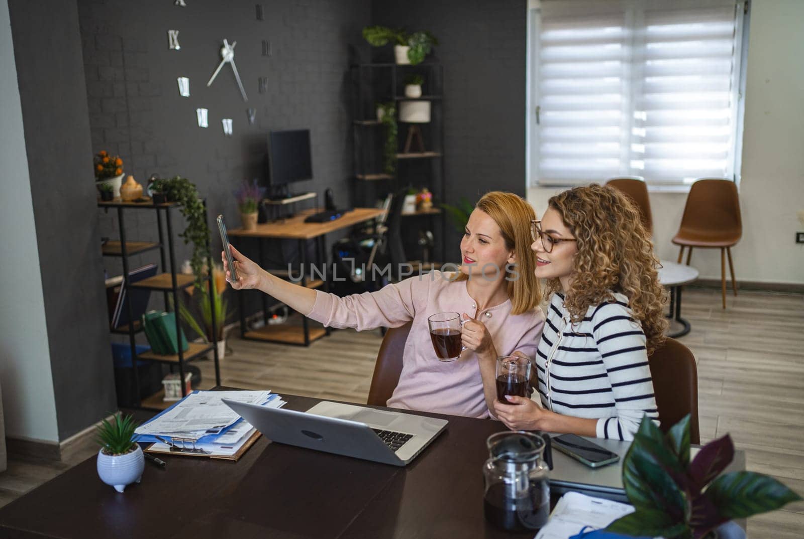 Two female colleagues are sitting at the table with a cup of coffee and taking a selfie.