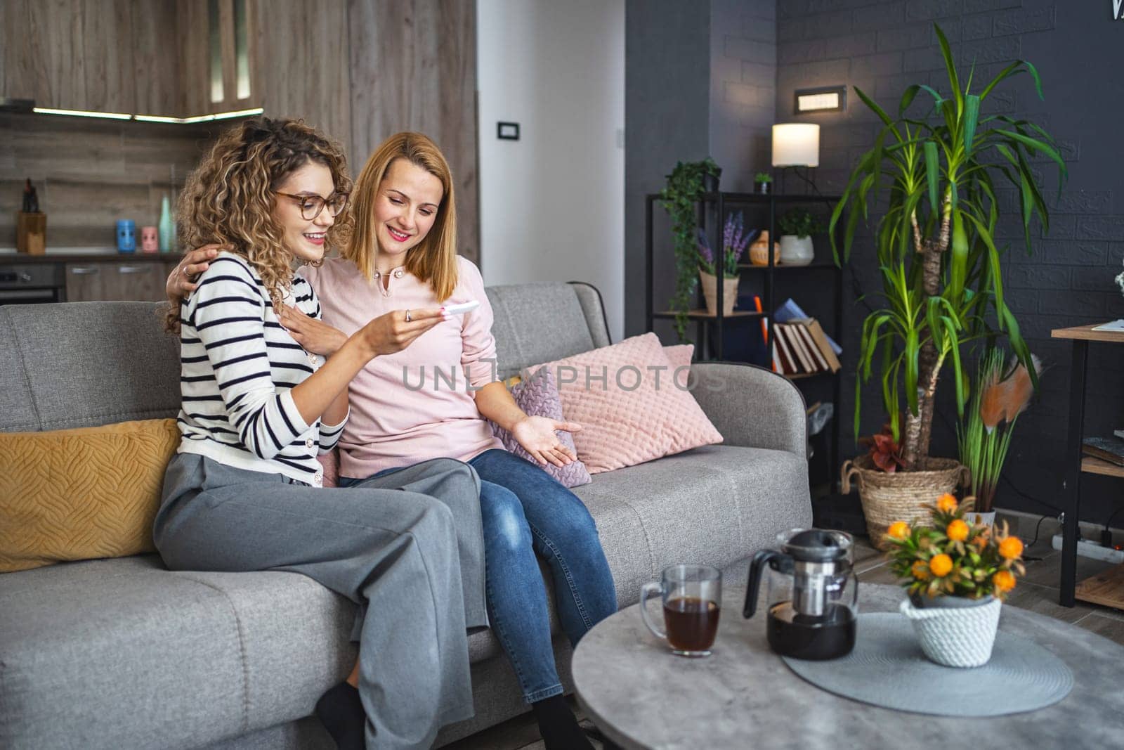 Two female friends are sitting on the couch in the living room, hugging and excited after getting their pregnancy test results.