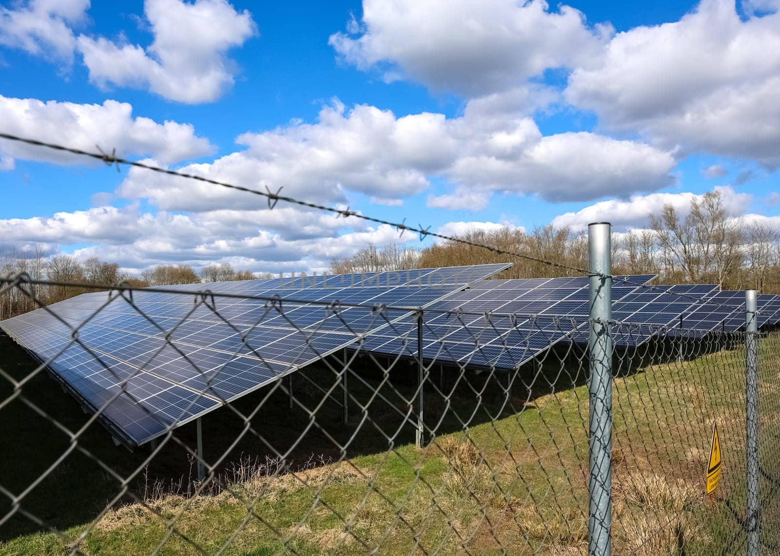 Photovoltaic panels generating renewable energy behind a barbed wire fence, under a cloudy blue sky