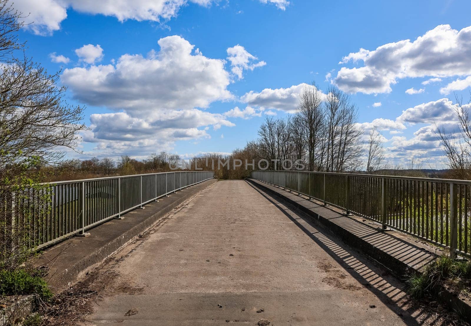 Empty narrow bridge with metal railing crossing a river in the countryside on a beautiful sunny spring day with some clouds