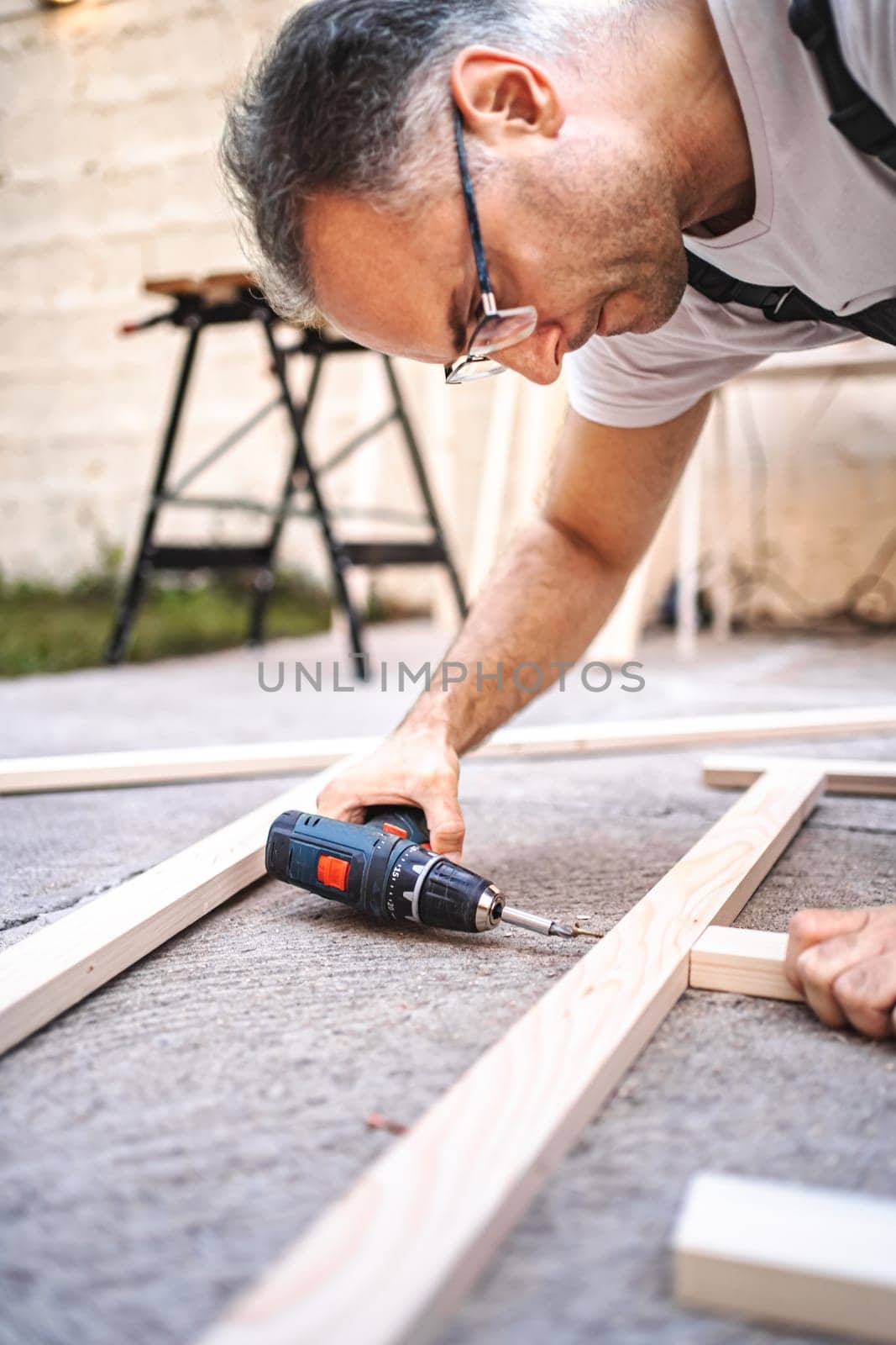 Mature male carpenter using electric screwdriver on a wooden plank outdoors.