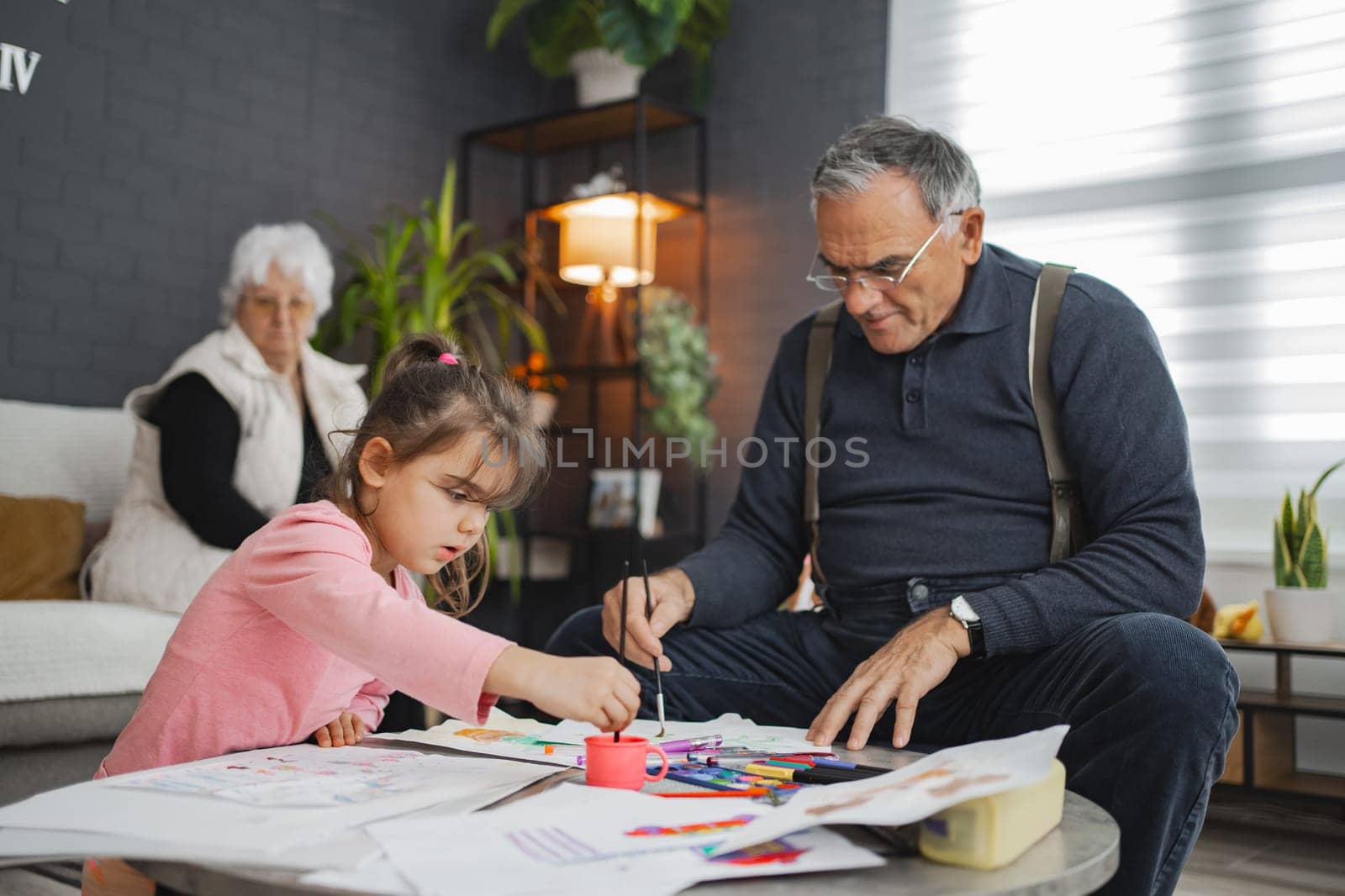 A young girl focuses on her artwork alongside her grandfather indoors.