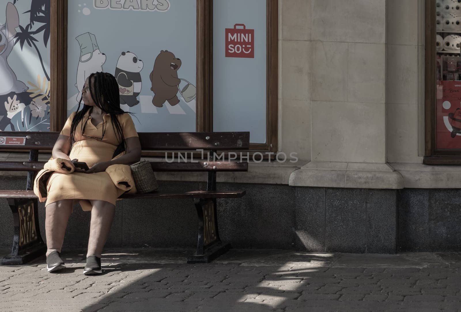 Windsor, UK - Apr 03, 2025 - African American women or Black women Wearing a yellow dress sitting on on a wooden bench in front of Miniso shop and holding a phone in hand. use it as your Wallpaper, Poster and Space for text, Selective focus.