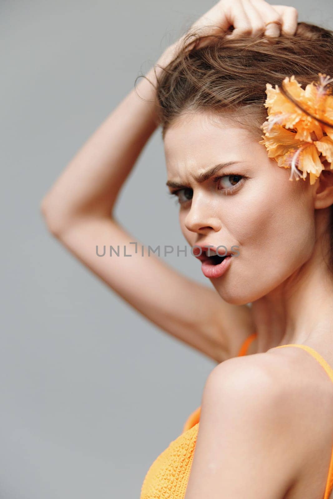 A fashionable young woman embodies summer energy with a playful expression, vibrant orange flower, and soft gray backdrop, ideal for fashion and emotional imagery.