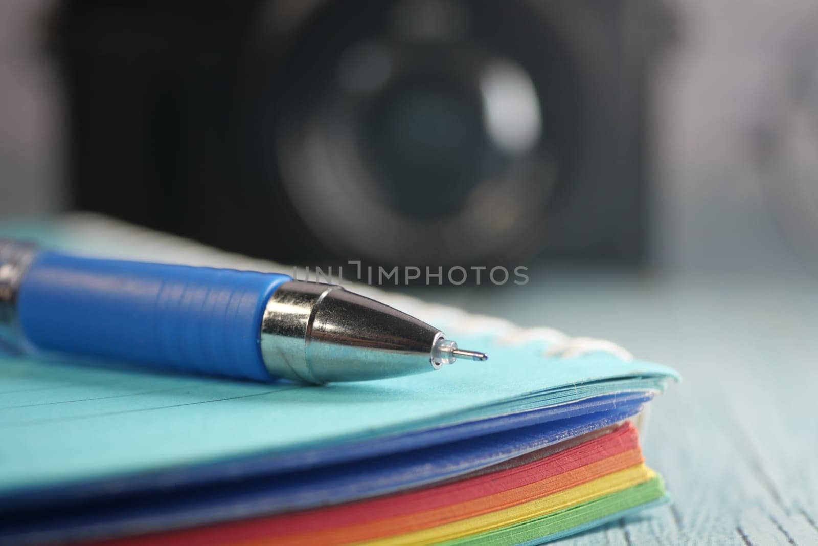 A blue pen rests on a stack of colorful paper near an unseen camera.