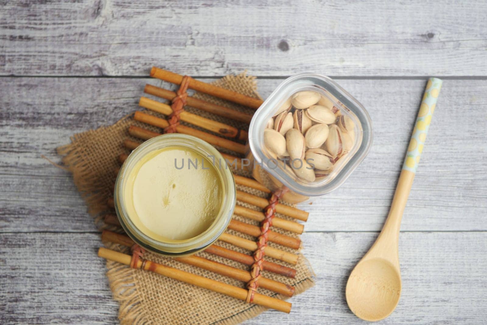 Nut spread sits next to pistachios and a spoon on a rustic surface.