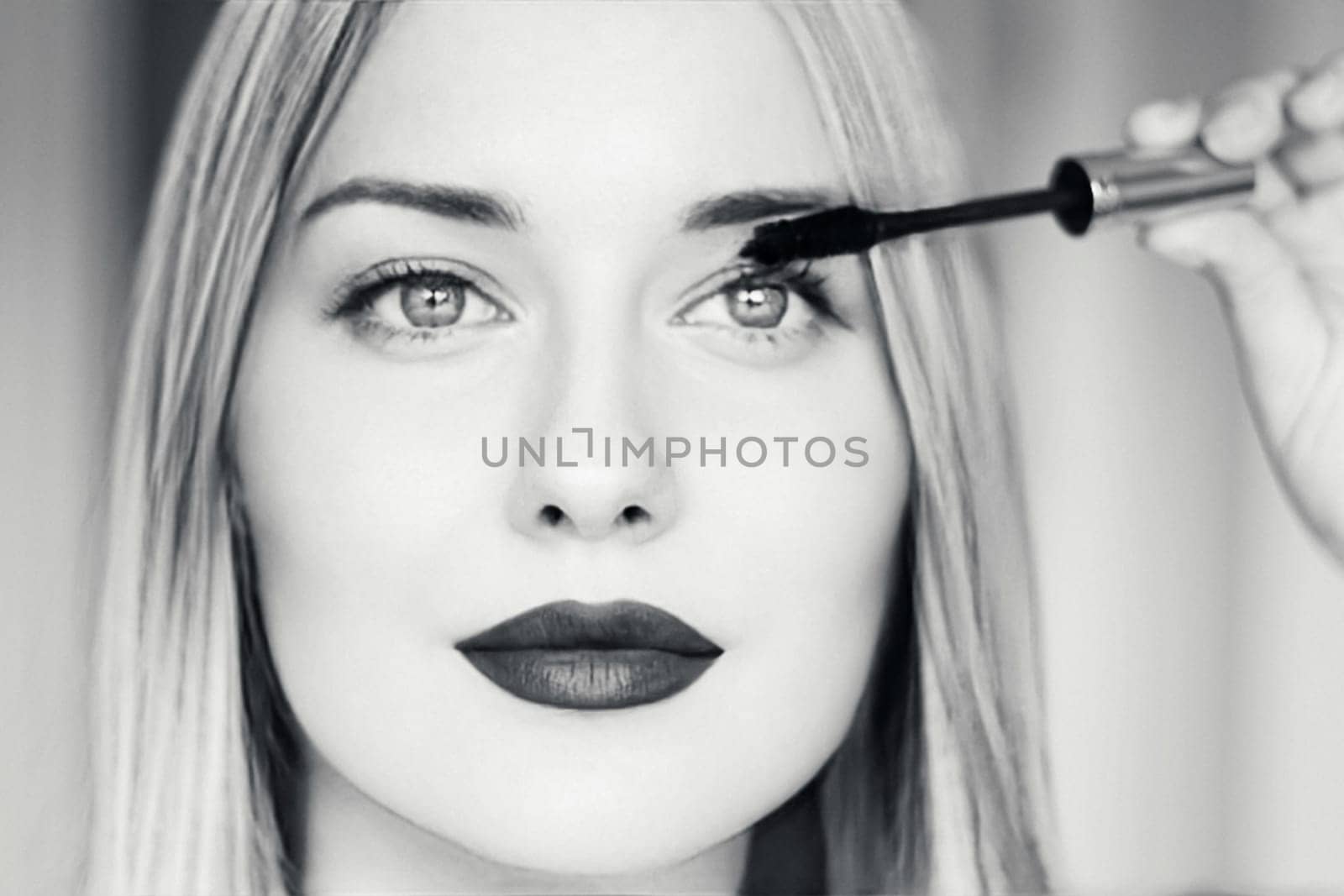 Close-up of a beautiful blonde woman applying mascara, with dark lipstick, bright eyes, and long straight hair. Black and white portrait for beauty, makeup, fashion, or cosmetic themes