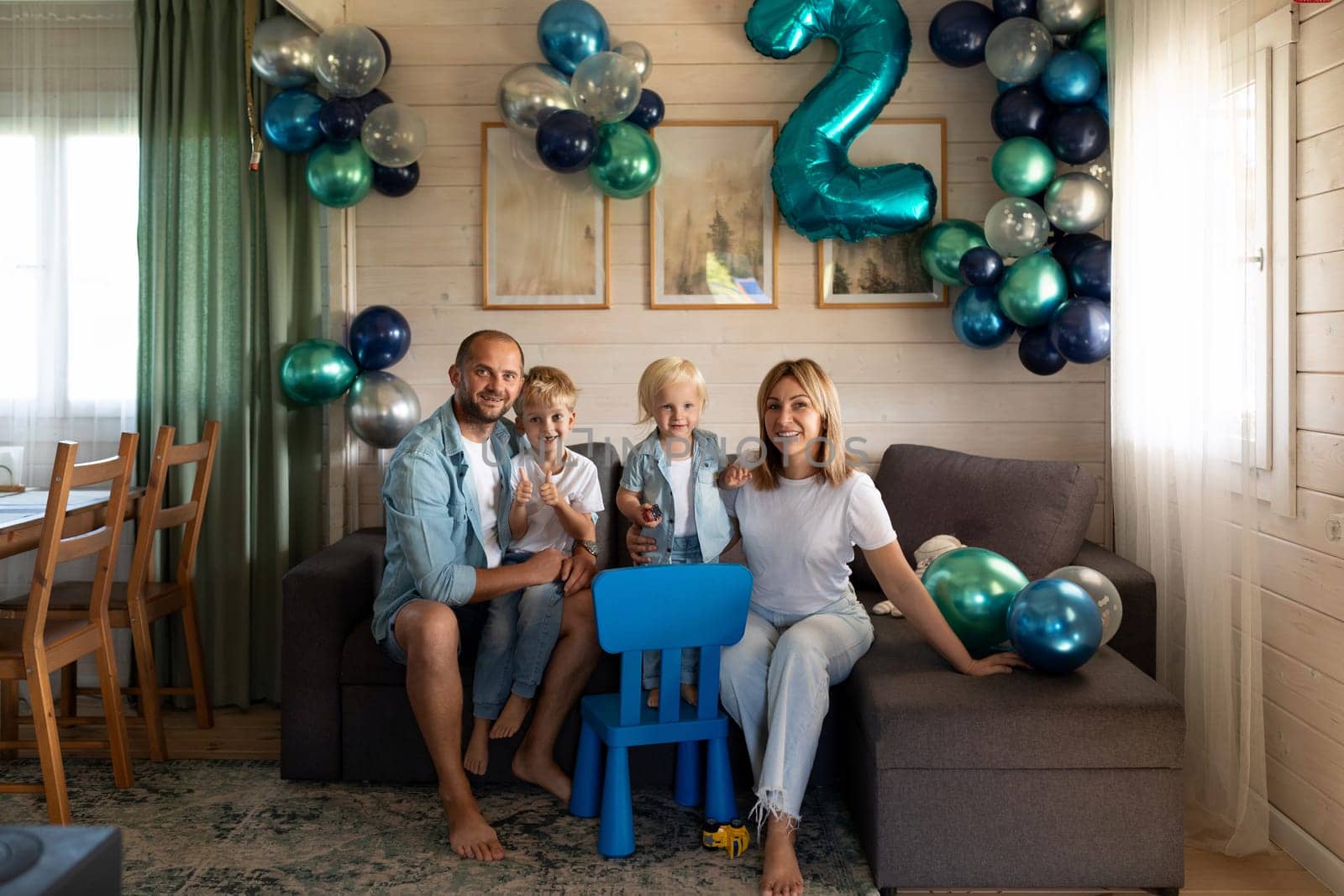 A family gathers in their comfortable living room to celebrate a child's second birthday. Colorful balloons and a blue chair add to the festive atmosphere while the family shares joyful moments.